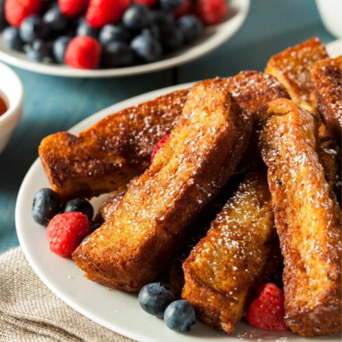 A plate filled with french toast sticks with blueberries and raspberries behind it