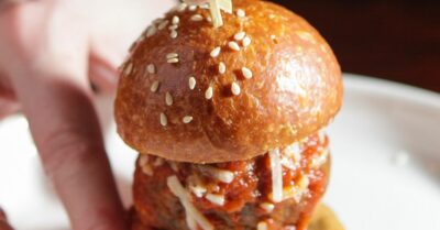 Hands holding a mini meatball slider with sesame seeds on the bun