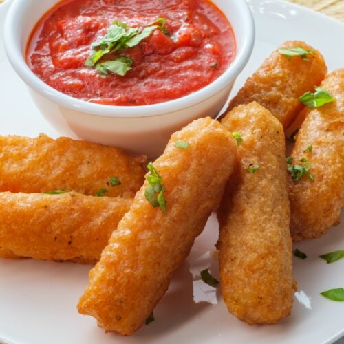 A plate filled with crispy and golden mozzarella sticks with marinara sauce on the side