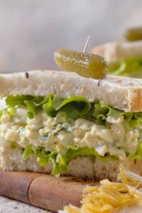 A masters egg salad sandwich on white bread with a pickle on top and lettuce coming out