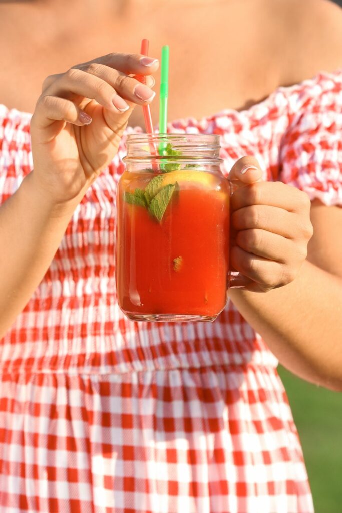 A woman in a red plaid dress drinking watermelon moonshine out of a mason jar with a green straw.