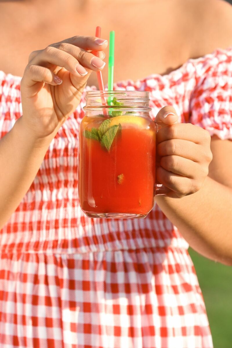 Watermelon Moonshine Recipe 2 A woman in a red plaid dress drinking watermelon moonshine out of a mason jar with a green straw.