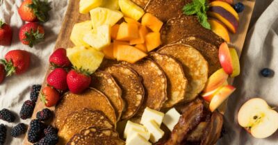 A wooden board filled with breakfast charcuterie ingredients like pancakes, egg and fruit.