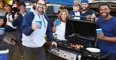 Friends around a grill smiling at a football tailgate party.