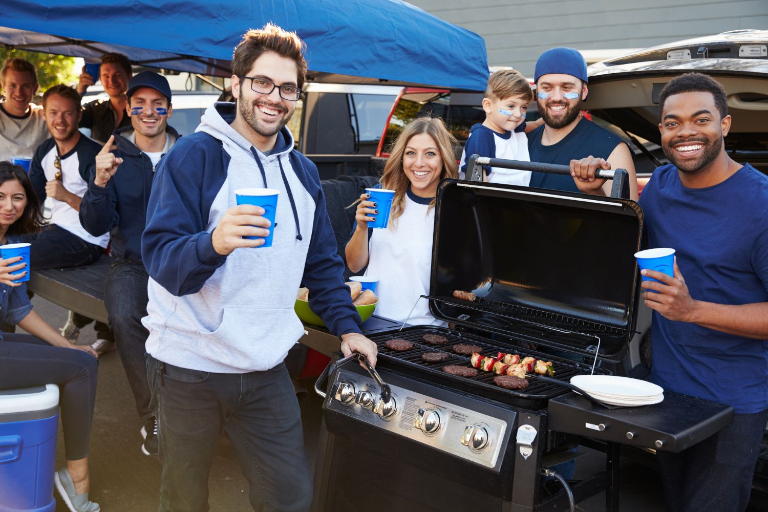 Friends around a grill smiling at a football tailgate party.