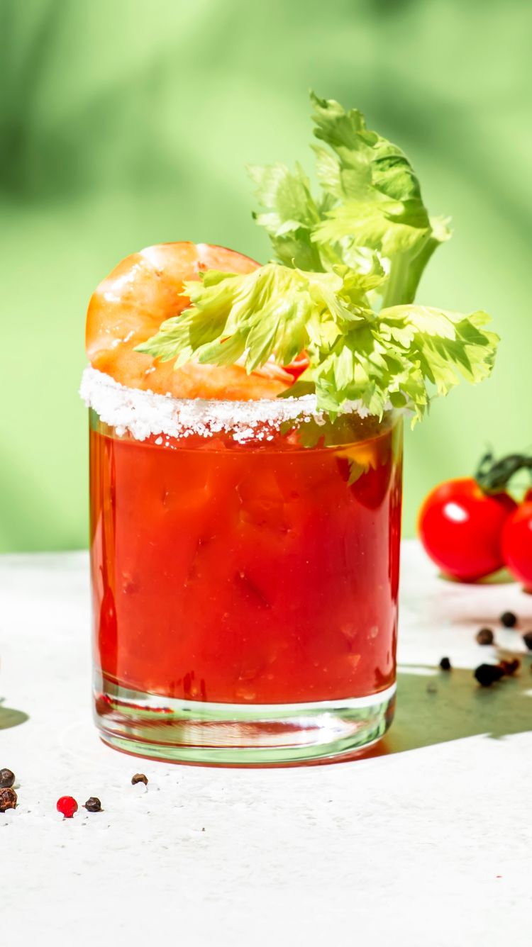 A caesar bloody mary in a glass with a salt rim and celery and shrimp in the glass.