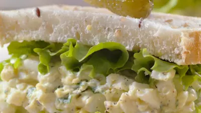 A masters egg salad sandwich on white bread with a pickle on top and lettuce coming out
