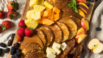A wooden board filled with breakfast charcuterie ingredients like pancakes, egg and fruit.